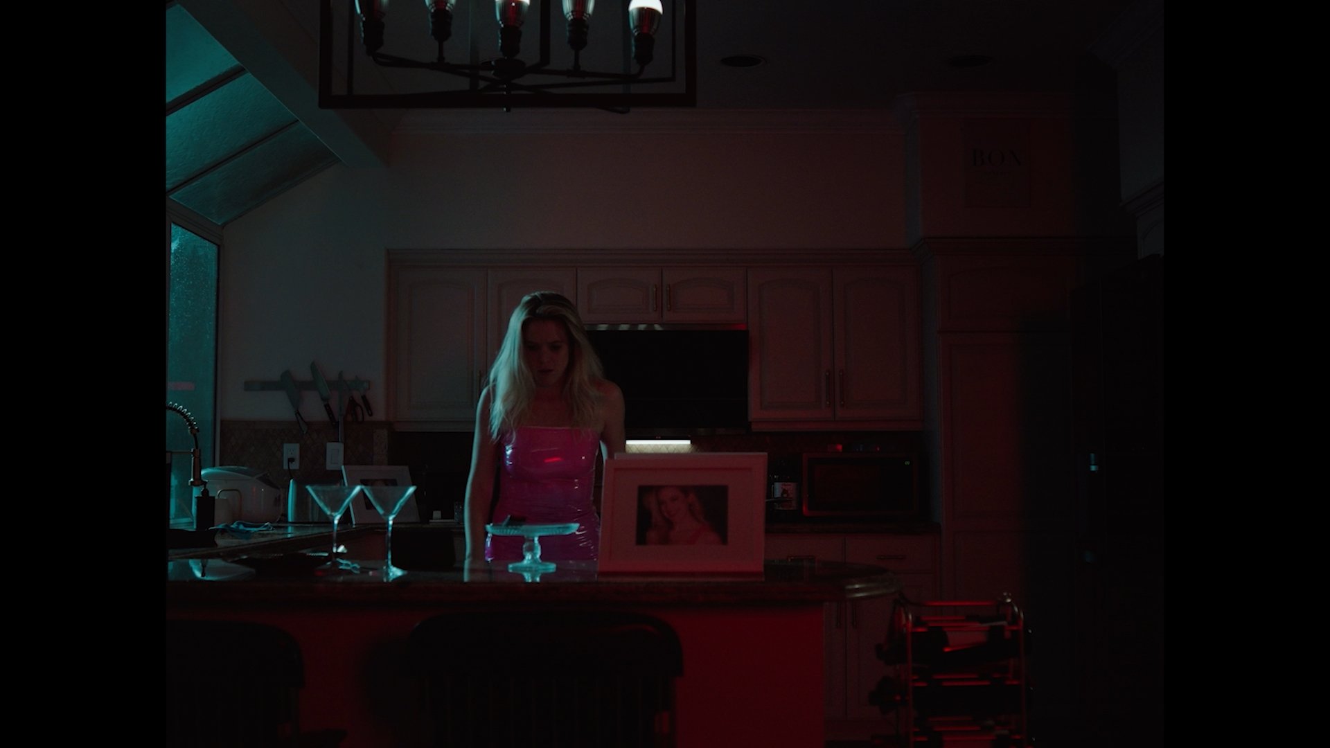A woman with blonde hair in a pink dress standing behind a kitchen counter at night, with two martini glasses and a framed photo on the counter, via dim lighting.