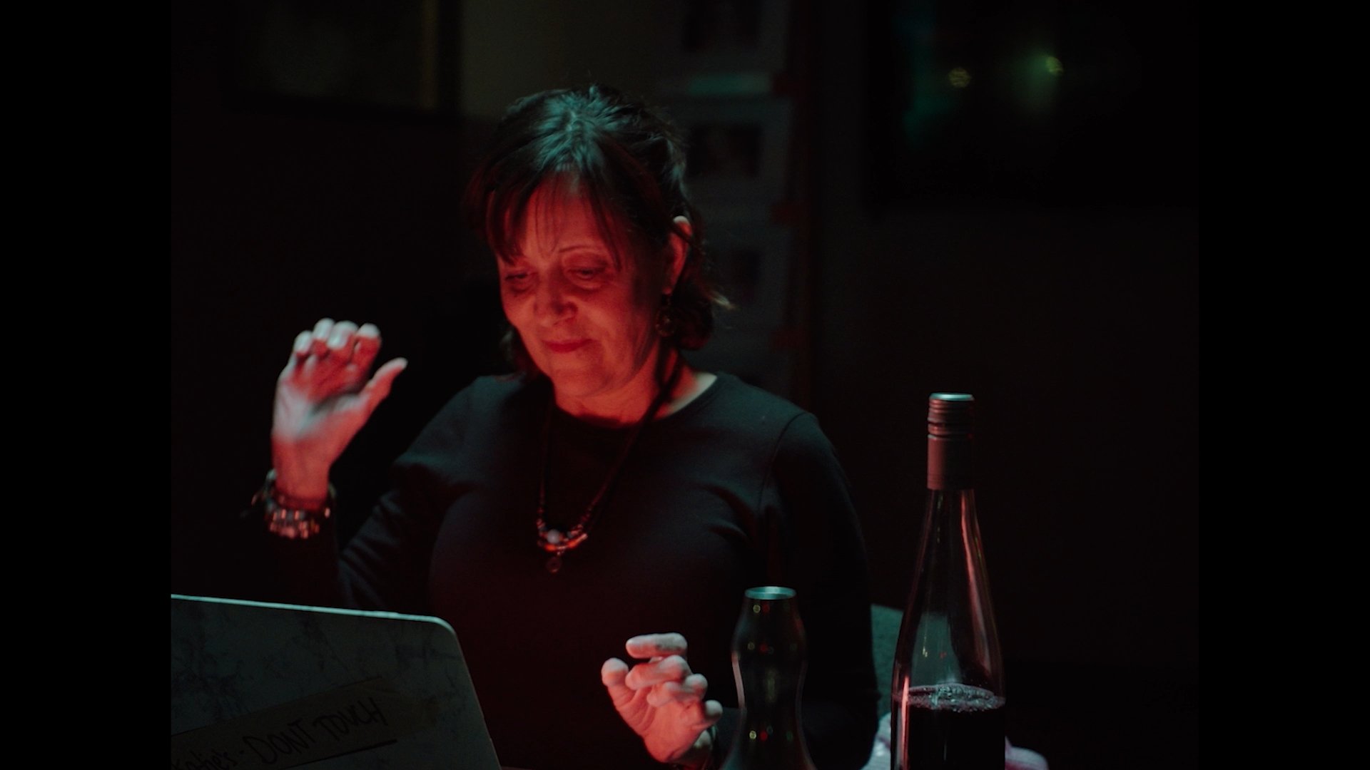 A woman with short dark hair, wearing a black shirt and colorful jewelry, appears to be in a dimly lit setting, possibly a bar or restaurant, illuminated by red lighting. She is looking at her phone with a slight smile, with a bottle and glass on the